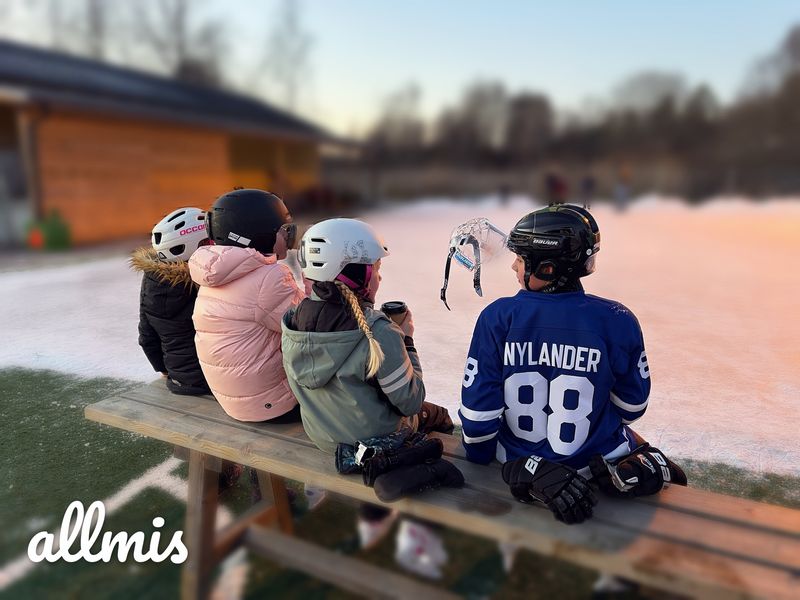 Fyra barn med hjälm och skridskor sitter på en bänk vid isbanan, med Allmis-logotypen